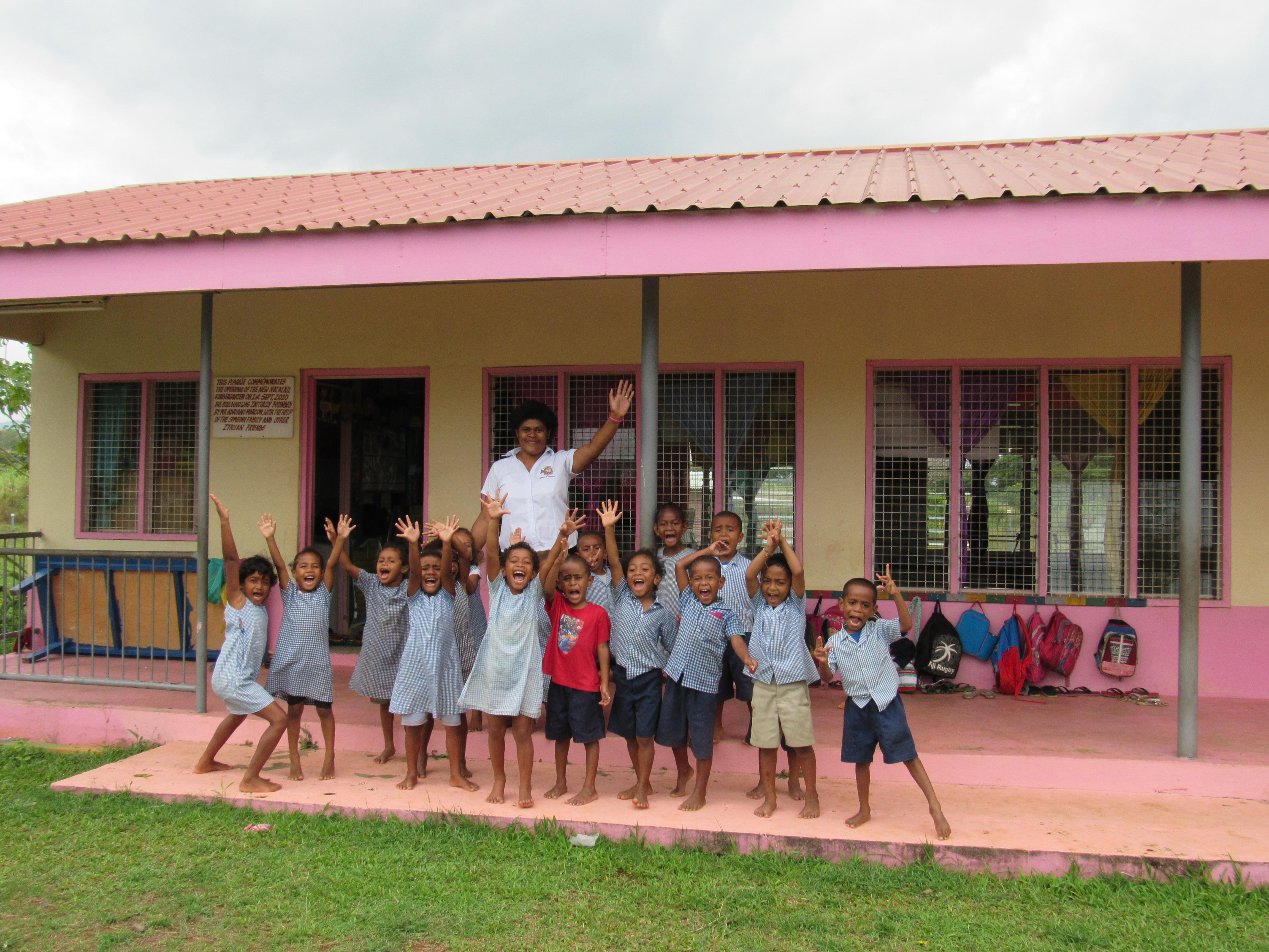 Kindergarten children wave with a teacher outside a school in Fiji where Projects Abroad Childcare volunteers are placed. A group of children pose for a photo with their teacher outside a kindergarten at one of our Childcare volunteer placements in Fiji.